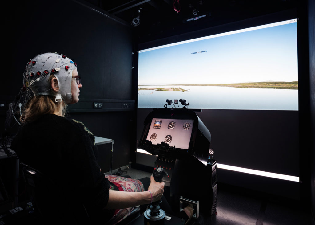 Experimental setup that shows a participant involved in a steering task (here a flight task). To measure the EEG, the participant is wearing an electrode cap (© Berthold Steinhilber / Max-Planck-Institut für biologische Kybernetik).