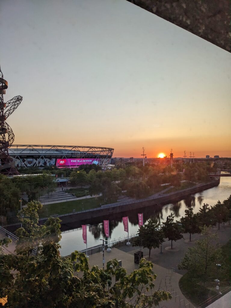 Upright photo showing the area in front of One Pool Street London with pink flags, trees and the waterworks river. Further, the London stadium is in the background as well as the setting sun.