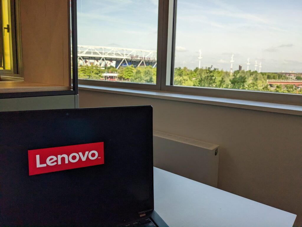 Part of a Laptop screen showing the Lenovo Logo while starting up in the foreground, in the background there are windows revealing a partial view on the London stadium.